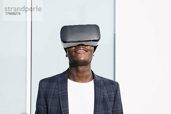 Lächelnder junger Mann mit VR-Brille vor der Wand