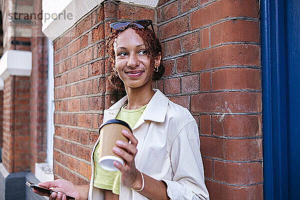 Lächelnde Frau hält Einwegkaffeetasse und Mobiltelefon an der Wand