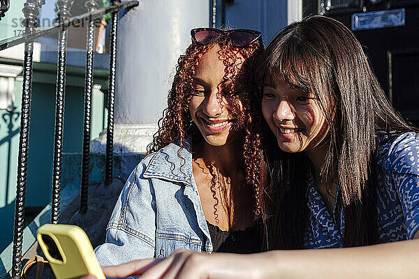 Lächelnde junge Frau mit Freundin  die an einem sonnigen Tag ihr Smartphone benutzt
