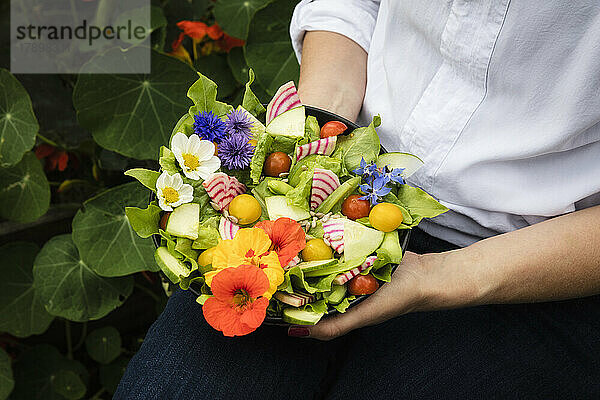 Mittelteil einer Frau  die eine Schüssel mit veganem Salat mit Gemüse und essbaren Blumen hält