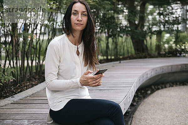 Lächelnde Geschäftsfrau mit Smartphone sitzt im Büropark