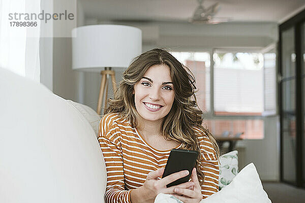 Porträt einer lächelnden jungen Frau mit Mobiltelefon auf dem heimischen Sofa