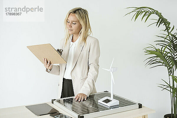 Frau hält Klemmbrett im Büro mit Solarpanel und Windturbinenmodell auf dem Schreibtisch