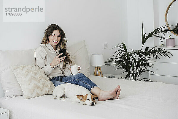 Junge Frau mit Kaffeetasse und Telefon sitzt zu Hause neben Hund im Bett