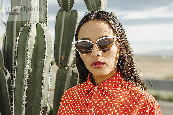 Selbstbewusste junge Frau mit Sonnenbrille steht vor einer Kaktuspflanze