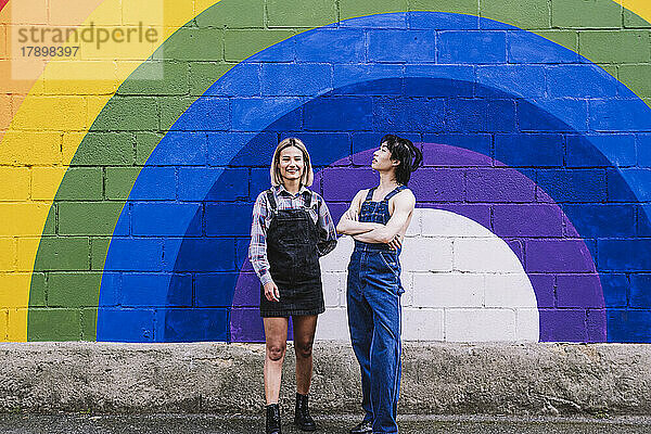 Glückliche junge Frau steht mit einer Freundin vor einem an die Wand gemalten Regenbogen