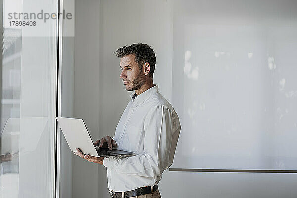Nachdenklicher Geschäftsmann mit Laptop und Blick durch das Fenster im Büro