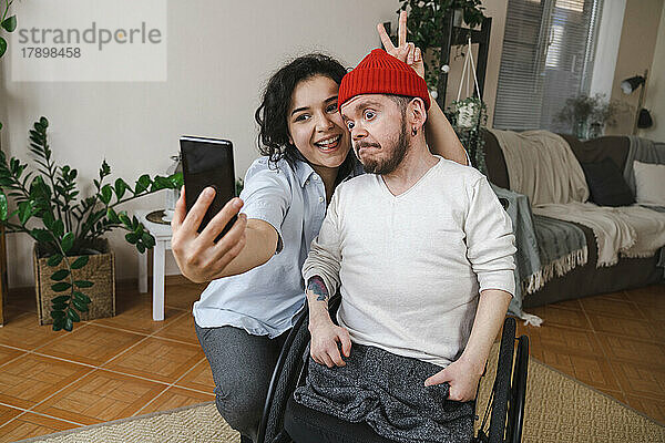 Glückliche Frau mit Mann im Rollstuhl  der zu Hause ein Selfie mit dem Handy macht
