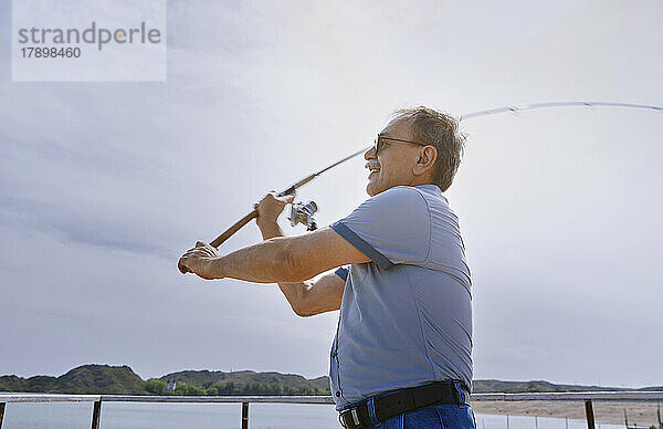 Älterer Mann fischt an einem sonnigen Tag mit der Angelrute auf dem Seeweg