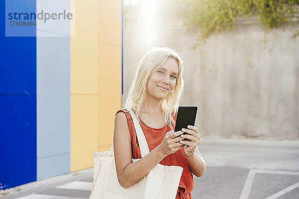 Lächelnde junge Frau mit blonden Haaren hält Smartphone in der Hand