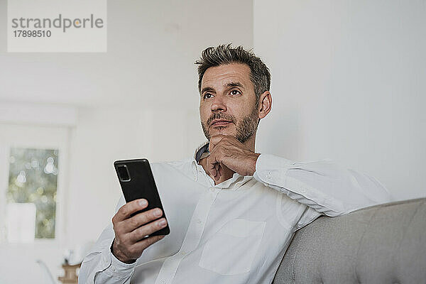 Nachdenklicher Geschäftsmann hält Smartphone auf dem Sofa im Büro