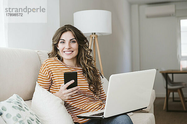 Lächelnde junge Frau mit Laptop und Mobiltelefon auf dem heimischen Sofa