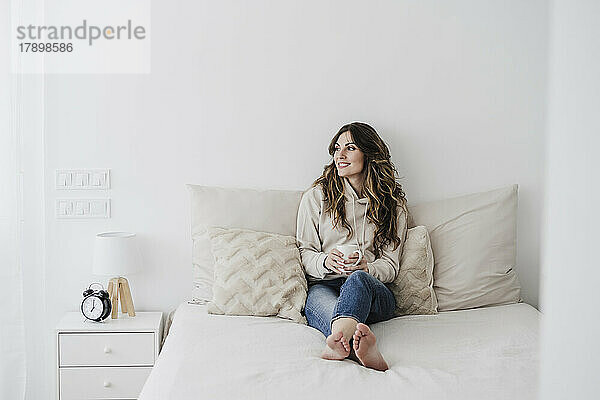 Lächelnde Frau mit Kaffeetasse sitzt zu Hause im Bett