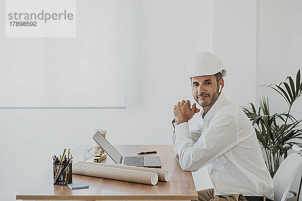 Ingenieur mit Schutzhelm sitzt mit Laptop am Schreibtisch im Büro