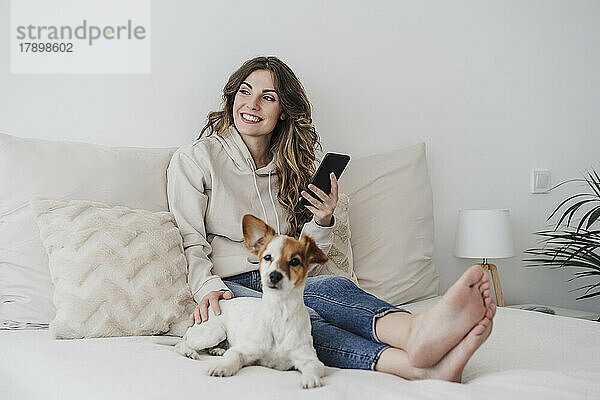 Lächelnde Frau mit Handy sitzt neben Hund im Schlafzimmer
