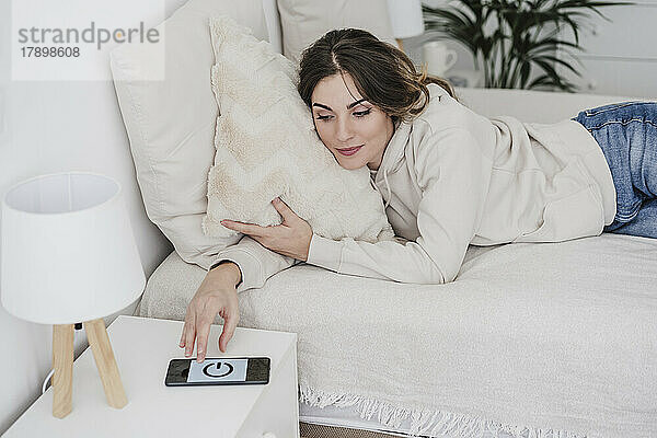 Junge Frau liegt im Bett und schaltet zu Hause den Smartphone-Wecker aus