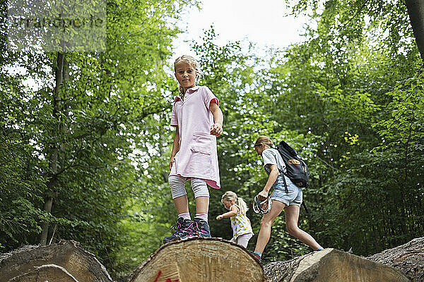 Lächelndes Mädchen steht auf einem Baumstamm und Mutter und Tochter gehen im Wald spazieren