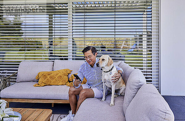 Smiling man using smart phone sitting with dog on sofa