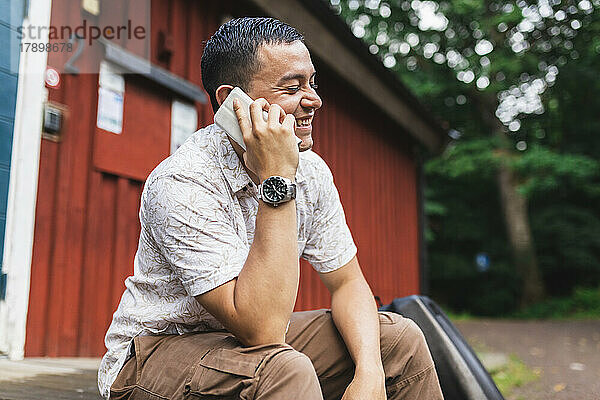 Lächelnder Mann mit geschlossenen Augen  der vor dem Haus sitzt und auf dem Smartphone spricht
