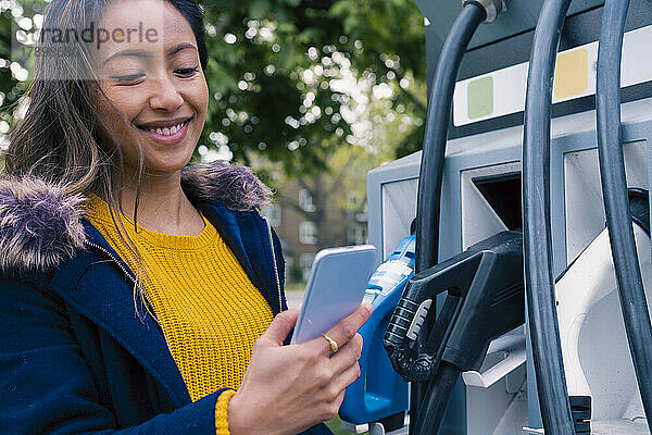 Lächelnde junge Frau benutzt Smartphone an der Ladestation für Elektrofahrzeuge