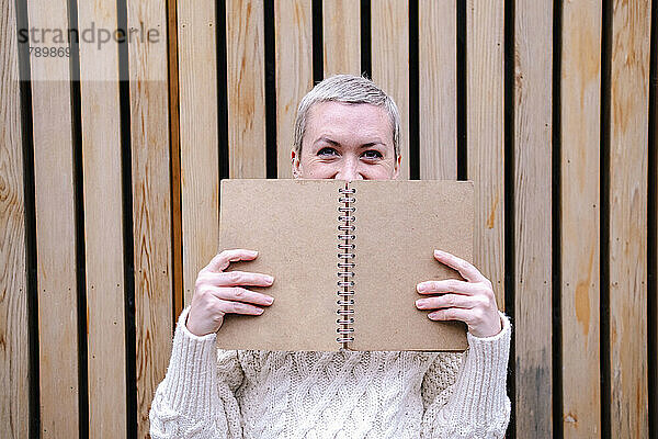 Frau versteckt Gesicht hinter offenem Buch vor Wand