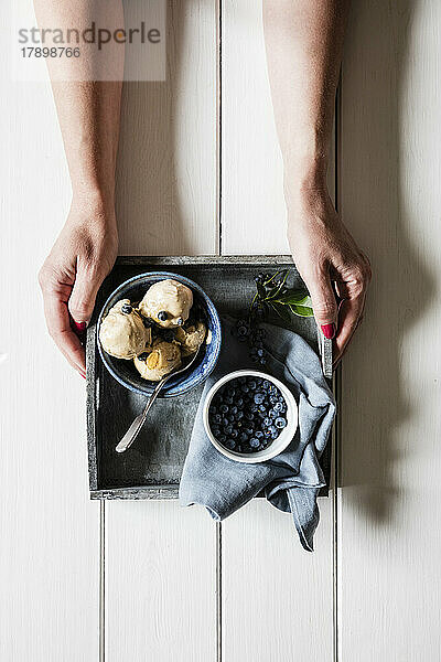 Arms of woman picking up tray with fresh blueberries and bowl of homemade peanut ice cream