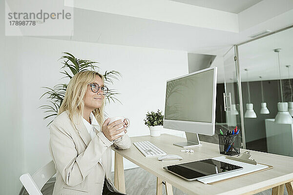 Geschäftsfrau sitzt am Schreibtisch im Büro und hält eine Tasse Kaffee in der Hand