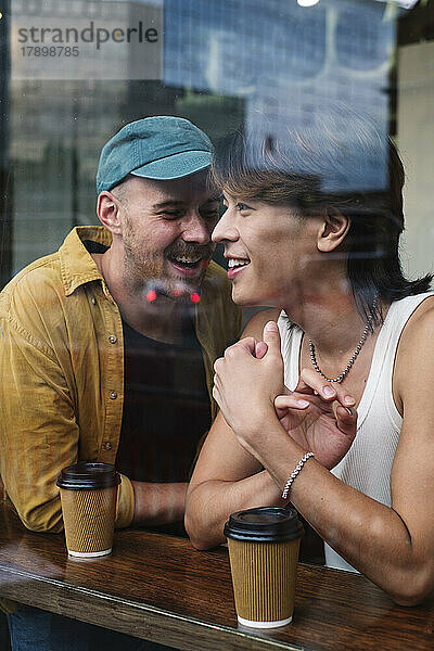 Lächelndes schwules Paar mit Kaffeetassen  gesehen durch das Fenster des Cafés