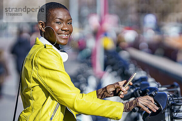 Junge Frau in Lederjacke hält Smartphone an Fahrradstation