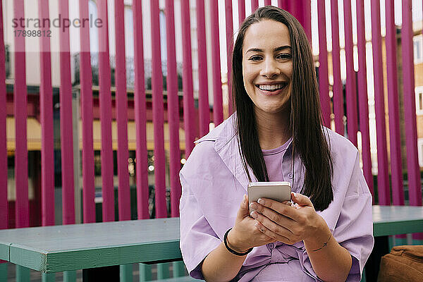 Glückliche junge Frau mit Smartphone sitzt auf Bank