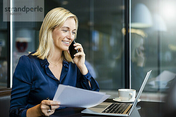 Glückliche Geschäftsfrau  die im Café auf dem Smartphone spricht und auf den Laptop schaut