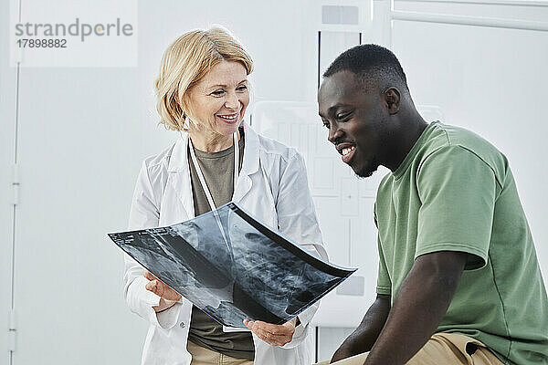 Lächelnder Arzt mit Röntgenbild  der den Patienten in der Klinik betrachtet