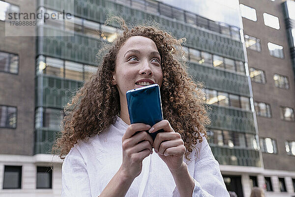 Nachdenkliche Frau mit lockigem Haar hält Smartphone vor Gebäude