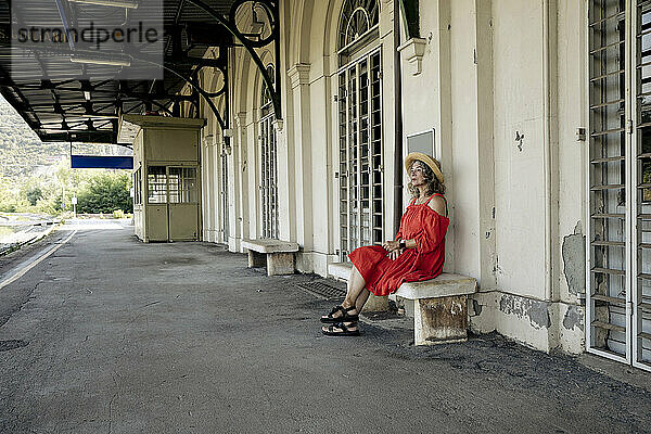Ältere Frau im roten Kleid sitzt auf einer Bank am Bahnhof