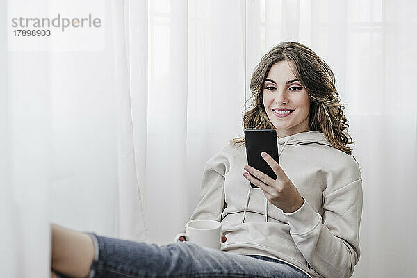 Lächelnde Frau mit Tasse und Handy zu Hause