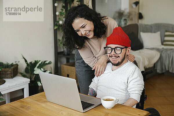 Lächelnde Frau steht neben ihrem Freund im Rollstuhl und schaut auf den Laptop