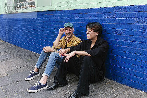 Smiling gay couple sitting on sidewalk sitting in front of building
