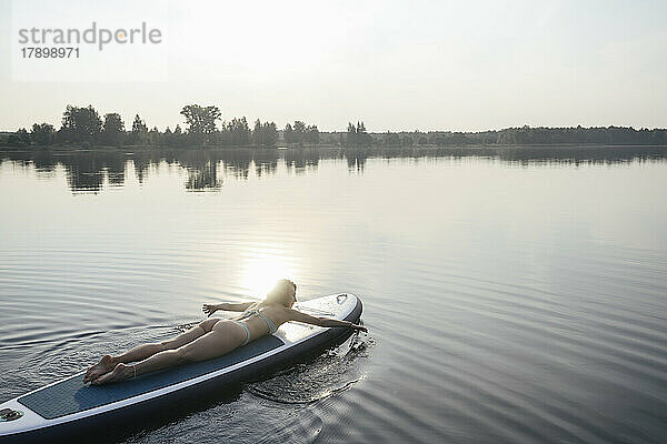 Frau paddelt mit der Hand und liegt bei Sonnenuntergang auf dem Paddleboard