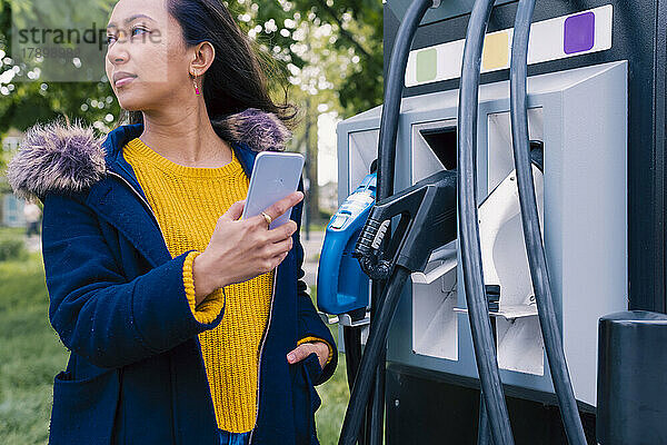 Junge Frau mit Mobiltelefon steht an einer Elektrofahrzeugstation