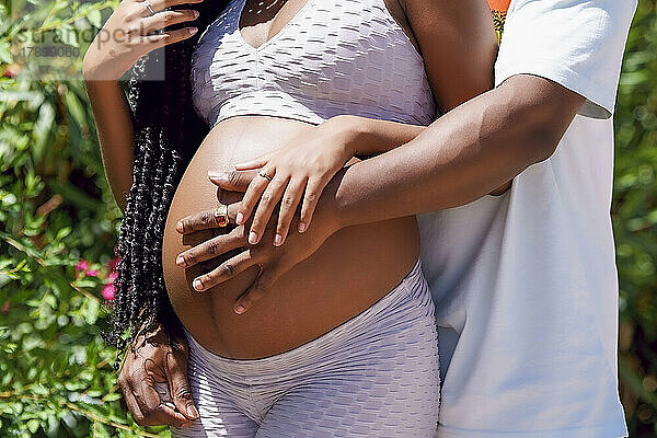 Mann umarmt den Bauch einer schwangeren Frau an einem sonnigen Tag