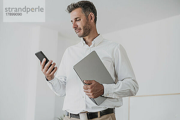 Lächelnder Geschäftsmann mit Laptop und Blick auf sein Mobiltelefon im Büro