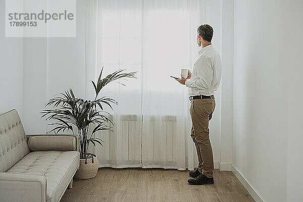 Geschäftsmann hält Mobiltelefon und Kaffeetasse in der Hand und schaut durch das Fenster im Büro