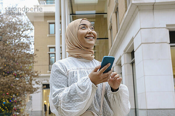 Glückliche junge Frau mit Smartphone steht vor dem Gebäude