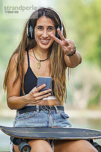 Glückliche junge Frau mit Smartphone zeigt Friedenszeichen-Geste