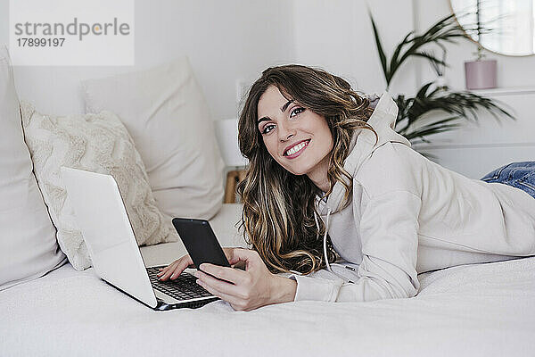 Glückliche Frau mit Laptop und Telefon  die zu Hause im Bett liegt