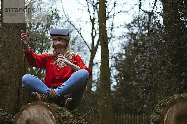 Junge Frau mit VR-Brille sitzt mit gekreuzten Beinen auf einem Baumstamm im Wald