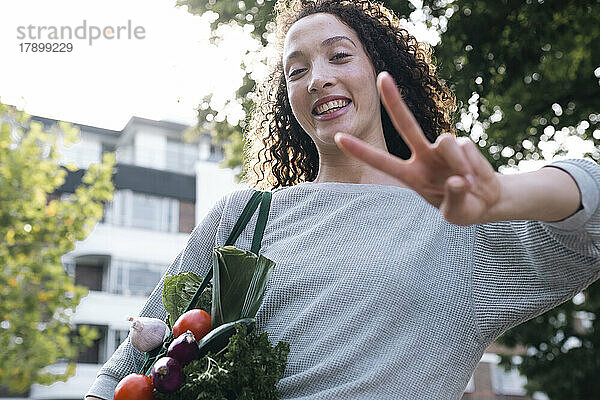 Fröhliche Frau mit einer Tüte Gemüse gestikuliert mit einem Friedenszeichen