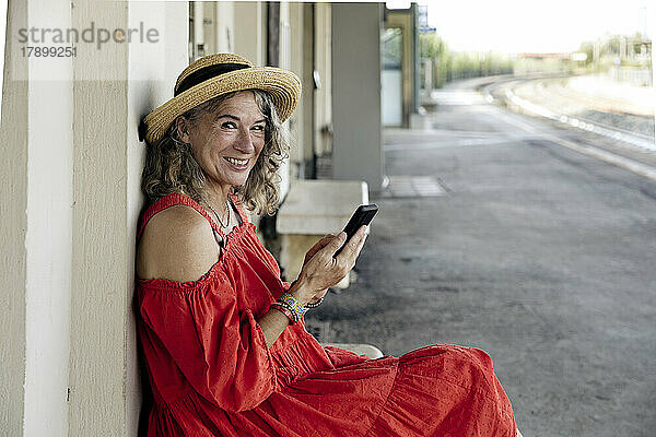 Lächelnde ältere Frau mit Smartphone sitzt am Bahnhof