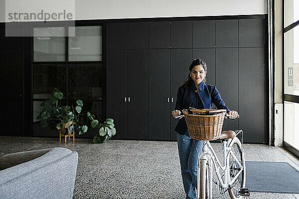 Lächelnde junge Geschäftsfrau mit Fahrrad steht in der Bürolobby