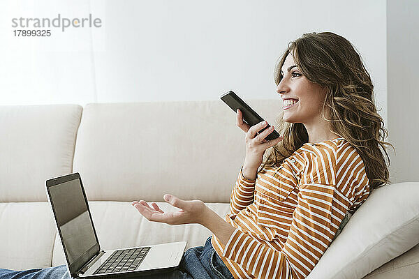 Junge Frau mit Laptop nutzt Mobiltelefon für eine Voicemail auf dem heimischen Sofa
